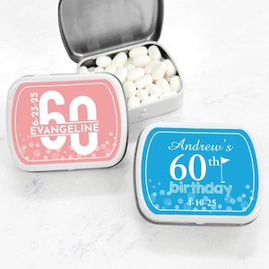 May include: Two small, rectangular mint tins with personalized labels. The tin on the left is pink with white polka dots and says "6-23-95 Evangeline 60". The tin on the right is blue with white polka dots and says "Andrew's 60th birthday 4-10-25".