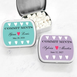 May include: Two small, rectangular mint tins with personalized labels. The tins are silver with white lids. The labels are teal and purple with white hearts and the text "COMMIT MINTS" in black. The teal tin has the names "Gina" and "Louis" with a pink heart and the date "May 20, 2026". The purple tin has the names "Sylvia" and "Martin" with a pink heart and the date "June 12, 2027".