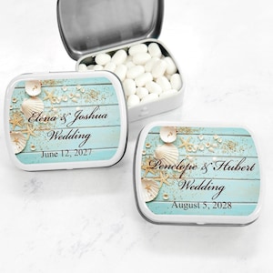 May include: Two small, white metal tins with blue and white labels. The labels feature a beach scene with seashells, starfish, and sand. The text on the labels reads "Elona & Joshua Wedding June 12, 2027" and "Penelope & Hubert Wedding August 5, 2028".