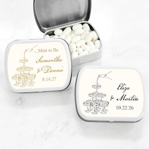 May include: Two white metal mint tins with personalized labels. The top tin has a gold design of a champagne tower and the text "Mint to Be Samantha & Donnie 8.14.27". The bottom tin has a black design of a champagne tower and the text "Eliza & Martin 10.22.26".