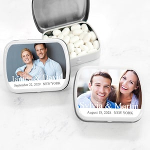 May include: Two silver mint tins with personalized labels featuring photos of couples and their names. The top tin says "Janice & David" and "September 22, 2029 NEW YORK". The bottom tin says "Sylvia & Martin" and "August 18, 2028 NEW YORK".