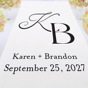 May include: White aisle runner with black calligraphy featuring the initials "KB" and the text "Karen + Brandon September 25, 2027". Two white and pink flower arrangements are on either side of the runner.