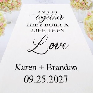 May include: White aisle runner with black text that reads "And so together they built a life they love Karen + Brandon 09.25.2027". The runner is decorated with white and pink flower arrangements.