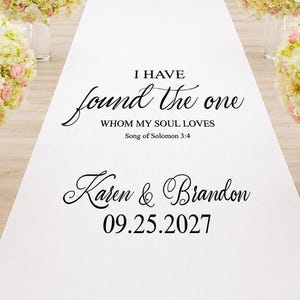 May include: A white wedding aisle runner featuring black printed text that reads I HAVE found the one WHOM MY SOUL LOVES Song of Solomon 3:4, followed by the names Karen & Brandon and the date 09.25.2027, flanked by four floral arrangements.