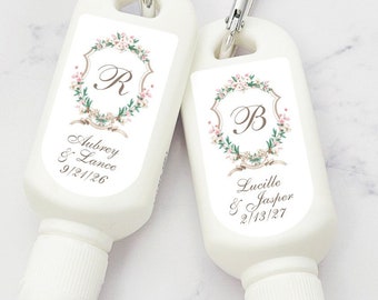 May include: Two white sunscreen bottles with floral designs and personalized wedding details. The bottles have a carabiner clip and are labeled with the couples' names and wedding dates: Aubrey & Lance 9/21/26 and Lucille & Jasper 2/13/27.
