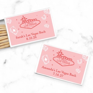 May include: Two pink matchboxes with white borders and red and white illustrations of a Las Vegas sign, playing cards, dice, and a horseshoe. The text on the boxes reads "Sarah's Las Vegas Bach 8.10.26" and "Amanda's Las Vegas Bach 7.14.27".