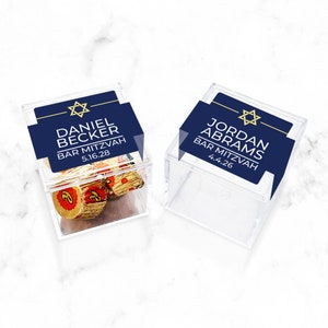 May include: Two clear plastic favor boxes with blue and gold labels. The labels have a Star of David and the text "Daniel Becker Bar Mitzvah 5.16.28" and "Jordan Abrams Bar Mitzvah 4.4.26".