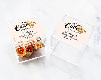 Op de afbeelding: Twee transparante plastic doosjes met deksels, elk met een kleine hoeveelheid chocoladebonbons. De doosjes hebben etiketten met de tekst "A Little Cutie is on the way" en de namen van de babyshowers, "Evelyn's Baby Shower 7.13.27" en "Leilanie's Baby Shower 5.10.26".