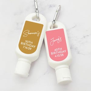 May include: Two small, white, plastic bottles with attached carabiners. The bottles have labels with personalized text. One label is gold with the text "Shannon's 50th Birthday 7.16.29". The other label is pink with the text "Jenny's 40th Birthday 4.12.28".