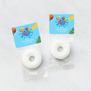 May include: Two favor bags with blue and white pool party themed labels. The labels have the text "Pool Party" and include a cartoon image of a pool float. The bags contain white round candies.