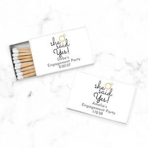 May include: Two rectangular white matchboxes with sliding trays. The top box is open, revealing wooden matches with white tips. Both boxes feature the text "she said Yes!" with a gold ring icon, plus personalized names and dates: "Debe's Engagement Party 8.20.27" and "Amelia's Engagement Party 1.12.28".