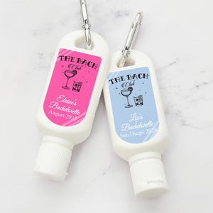 May include: Two small, white, travel-sized bottles of sunscreen with a carabiner clip. The bottles have personalized labels with a cocktail graphic and the text "THE BACH Club". One label is pink with the text "Elaine's Bachelorette August 2025" and the other is blue with the text "Lia's Bachelorette San Diego 2026".