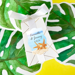 May include: White favor box with a gold geometric pattern and a white ribbon. A tag with a blue and white beach scene and the text "Carmella & Jeremy 6.18.24" is attached to the box.