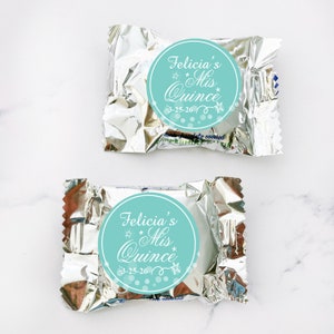 May include: Two silver foil-wrapped chocolate candies with round turquoise blue stickers. The stickers have white text that reads "Felicia's Quince" with stars and a date.