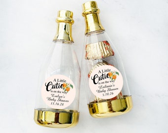 Op de afbeelding: Twee doorzichtige plastic champagneflessen met gouden doppen en etiketten waarop staat "A Little Cutie is on the way". Eén etiket zegt "Evelyn's Baby Shower 11.16.28" en de andere zegt "Leilanie's Baby Shower 5.10.26".