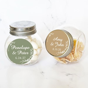 May include: Two small glass jars with silver lids and personalised labels. The jars are filled with sweets and have a green and brown floral design. The labels read "Penelope & Peter 6.24.27" and "Amy & John 8.10.26".
