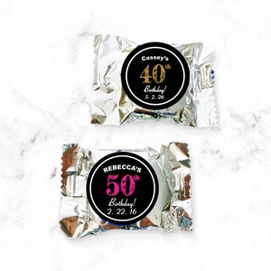 May include: Two silver foil-wrapped chocolate candies with personalized birthday stickers. The top sticker is black with gold text that reads "Cassey's 40th Birthday! 5.2.26". The bottom sticker is black with pink text that reads "REBECCA'S 50th Birthday! 2.22.16".
