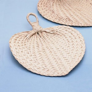 May include: Two woven straw fans, one is a round shape and the other is a heart shape. Both fans are a light beige color.