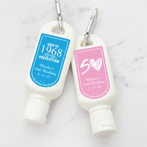 May include: Two small, white, plastic bottles with blue and pink labels. The blue label says "Aged to 1968 Vintage Perfection Charles's 50th Birthday 8-12-27". The pink label says "50 Meghan's 50th Birthday 2-15-26".