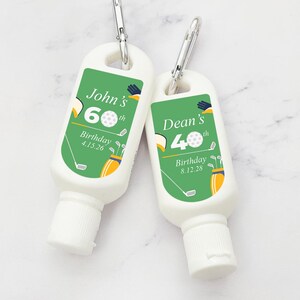 May include: Two green and white personalised sunscreen bottles with a golf theme. The bottles have a carabiner clip and say "John's 60th Birthday 4.15.26" and "Dean's 40th Birthday 8.12.28".