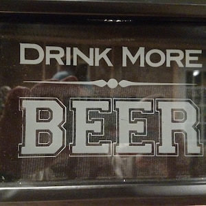 May include: Black framed sign with the words "DRINK MORE BEER" in white, bold, block letters. The sign is displayed on a wooden surface. The image is well-lit, highlighting the text.
