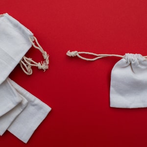 May include: A set of five white cotton drawstring bags on a red background. The bags are stacked on top of each other and have a natural cotton drawstring. There is a single white cotton drawstring bag on the right side of the image.