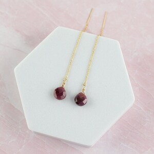 May include: Gold-coloured threader earrings with faceted, round, dark red gemstones. The earrings are displayed on a white, geometric stand against a pink marble background.