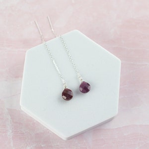 May include: A pair of silver threader earrings featuring faceted, teardrop-shaped dark pink gemstones. The earrings consist of a thin silver chain that passes through the ear, ending in a small, polished, deep magenta stone drop.