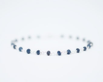 May include: A delicate necklace with small, round, dark blue beads strung on a fine silver chain. The beads are evenly spaced, creating a minimalist and elegant design. The necklace is set against a bright white background.