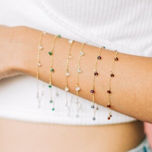 May include: A close-up of a person's arm wearing multiple delicate gold chain bracelets with small gemstone accents. The bracelets feature different colored gemstones, including green, white, and red.