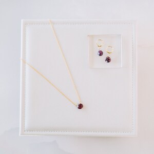 Op de afbeelding: Een gouden ketting met een enkele gefacetteerde donkerrode edelsteen hanger, vergezeld door een paar bijpassende gouden oorringen, elk met een kleine, gefacetteerde donkerrode edelsteen als bungelend element in de set.