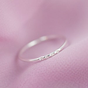 May include: A simple silver band ring with a textured design. The ring is on a pink fabric background.
