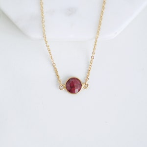 Puede incluir: Un delicado collar de oro con un colgante redondo de piedra preciosa roja oscura. El colgante es facetado y está engastado en un bisel de oro. La cadena es fina. El collar se muestra sobre una superficie blanca, con vegetación borrosa en el fondo.