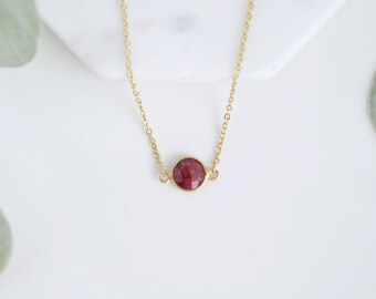 May include: A delicate gold necklace featuring a faceted, round, deep red gemstone pendant. The chain is fine and the pendant is set in a gold bezel. The necklace is displayed on a white surface with soft, blurred greenery in the background.