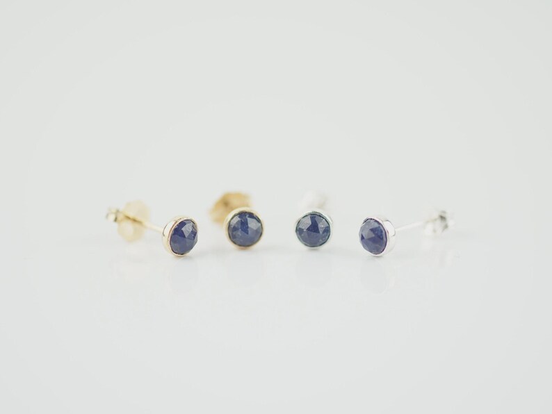 May include: Two pairs of stud earrings featuring round, faceted dark blue stones set in metal. One pair is set in a warm-toned yellow metal, while the other pair is set in a cool-toned silver-colored metal. Each earring has a post back closure.