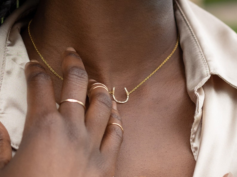 Puede incluir: Un collar de cadena de oro con un peque&ntilde;o colgante de herradura. El collar lo lleva una persona de piel oscura.