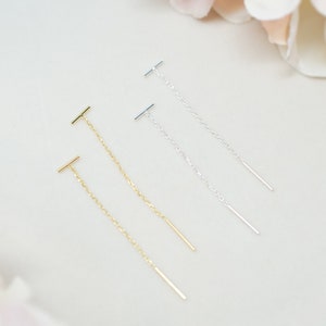 May include: Three pairs of gold and silver dangle earrings with a thin chain and a small bar at the end.