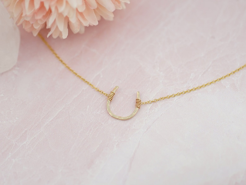 Puede incluir: Un collar de herradura de oro sobre un fondo rosa. La herradura est&aacute; hecha de metal martillado y tiene una cadena delicada.