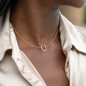 K&ouml;nnte beinhalten: Eine goldene Kettenkette mit einem kleinen Hufeisen-Anh&auml;nger. Die Kette wird von einer Person mit dunklen Haaren getragen.