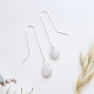 May include: Silver threader earrings with pear-shaped moonstone pendants. The earrings are suspended from fine chains, creating a minimalist design. The background is a soft white. The moonstones have an iridescent quality.
