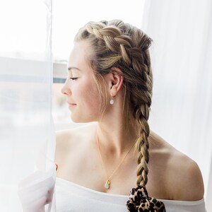 May include: A woman with blonde hair styled in a braid with a leopard print scarf tied around the braid. She is wearing a white off-the-shoulder top and a gold necklace with a white stone pendant.