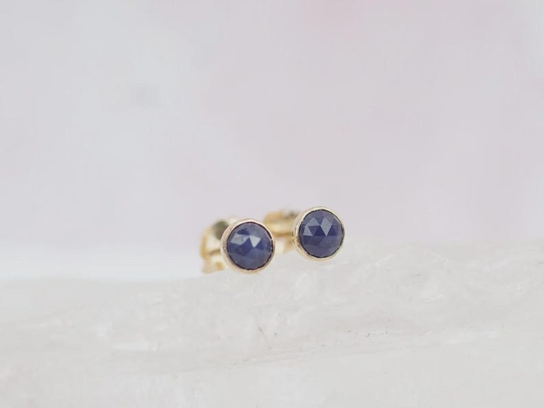May include: A pair of stud earrings featuring round, faceted dark blue sapphire stones set in polished gold bezels. The stones have a geometric, rose-cut surface texture that reflects light, and the earrings are secured with gold post backings.