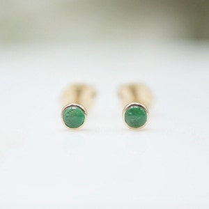 May include: A pair of small, round emerald green gemstone earrings with gold settings.