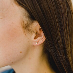 May include: A small, round, gold-toned stud earring featuring a clear, circular gemstone set in the center. The earring is worn in the earlobe, showcasing a simple, minimalist design with a polished metal finish and a clear, reflective stone.
