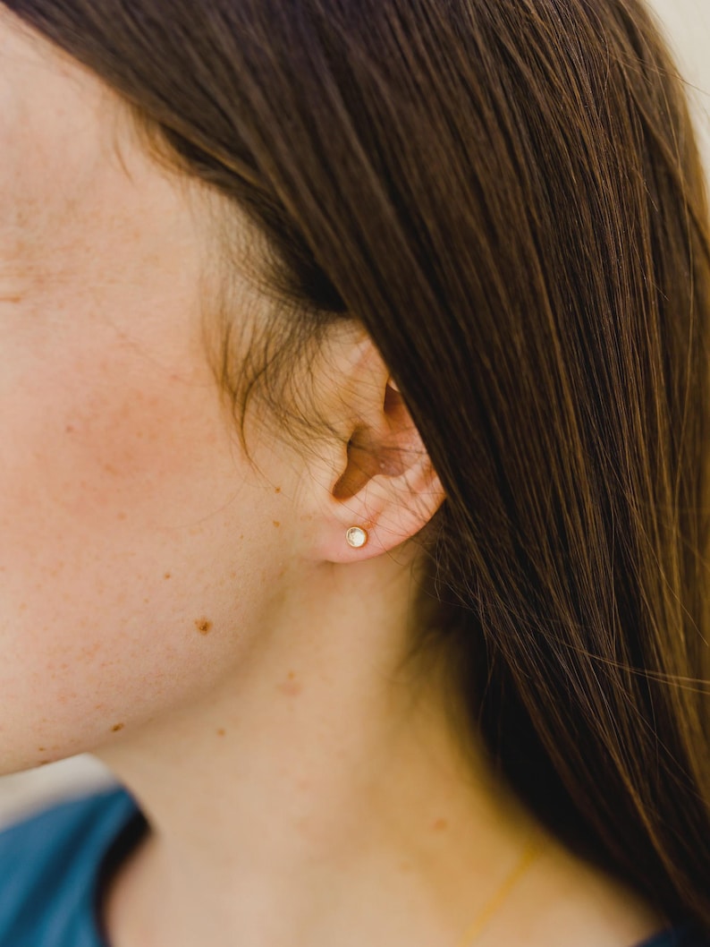 May include: A small, round, gold-toned stud earring featuring a clear, circular gemstone set in the center. The earring is worn in the earlobe, showcasing a simple, minimalist design with a polished metal finish and a clear, reflective stone.