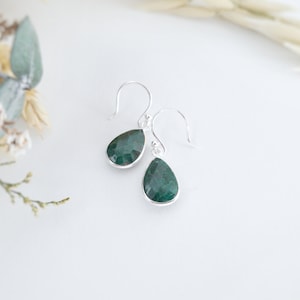 May include: A pair of silver teardrop earrings with faceted, deep green gemstones. The earrings have a simple hook design and are set against a white background, with soft floral accents in the upper left corner.