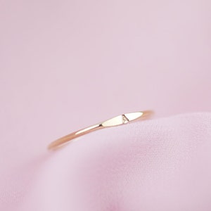 May include: A thin, polished gold-toned ring featuring a small, flat, horizontal bar section on the top. The center of this flat bar is engraved with the capital letter A. The ring has a smooth, reflective metallic finish throughout its band.