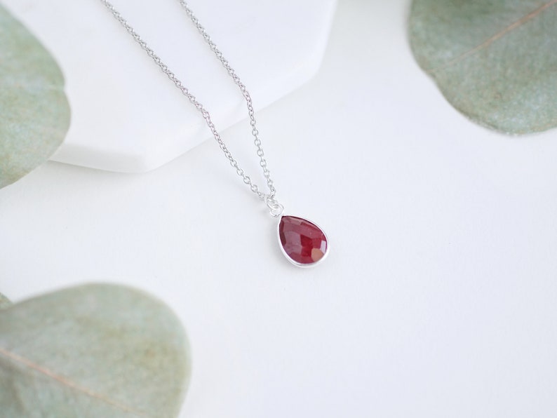 May include: A silver chain necklace with a teardrop-shaped pendant featuring a deep red gemstone.