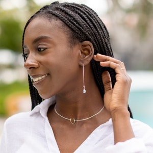 Puede incluir: Una mujer con largas trenzas negras lleva una camisa blanca y un collar de plata con un colgante de herradura. Lleva pendientes largos de plata con una peque&ntilde;a piedra azul en el extremo.