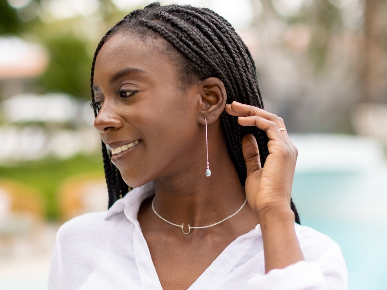 Puede incluir: Una mujer con largas trenzas negras lleva una camisa blanca y un collar de plata con un colgante de herradura. Lleva pendientes largos de plata con una peque&ntilde;a piedra azul en el extremo.